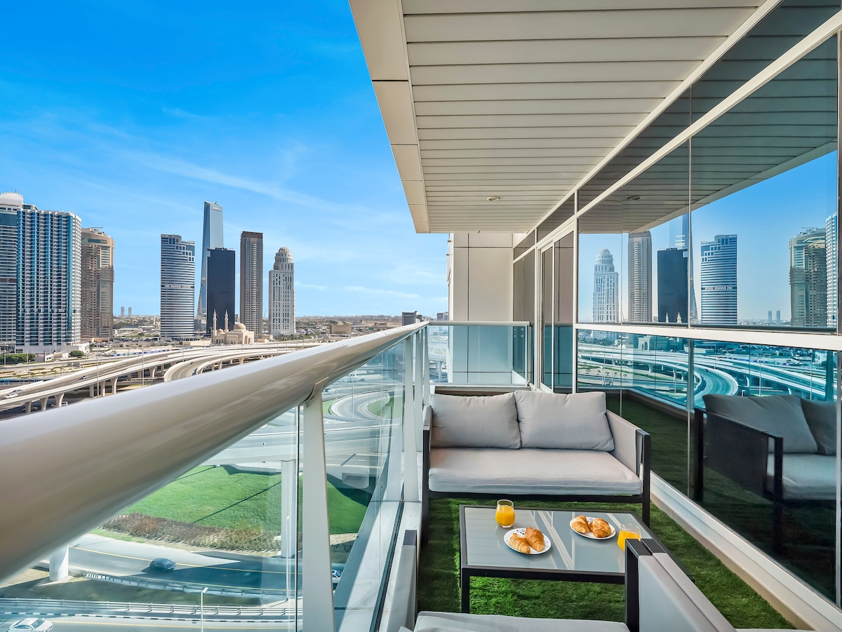 A balcony space features modern furniture, including a table with a light breakfast set of croissants and orange juice. Floor-to-ceiling glass railings provide unobstructed views of the cityscape, showcasing tall buildings under a clear blue sky.