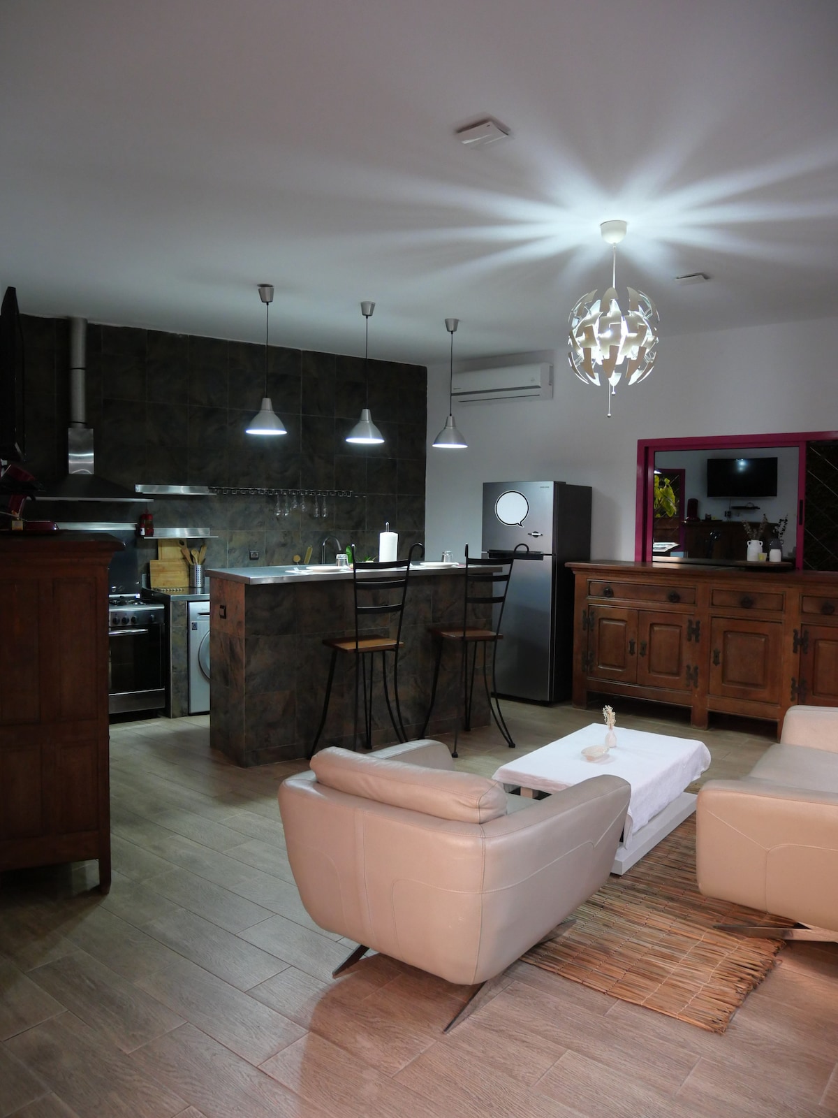 A modern kitchenette is visible, featuring a refrigerator and a sleek stovetop. Two bar stools are positioned at a counter, while a light fixture hangs above. A cozy seating area includes light-colored armchairs, with a low table resting on a textured rug beneath.