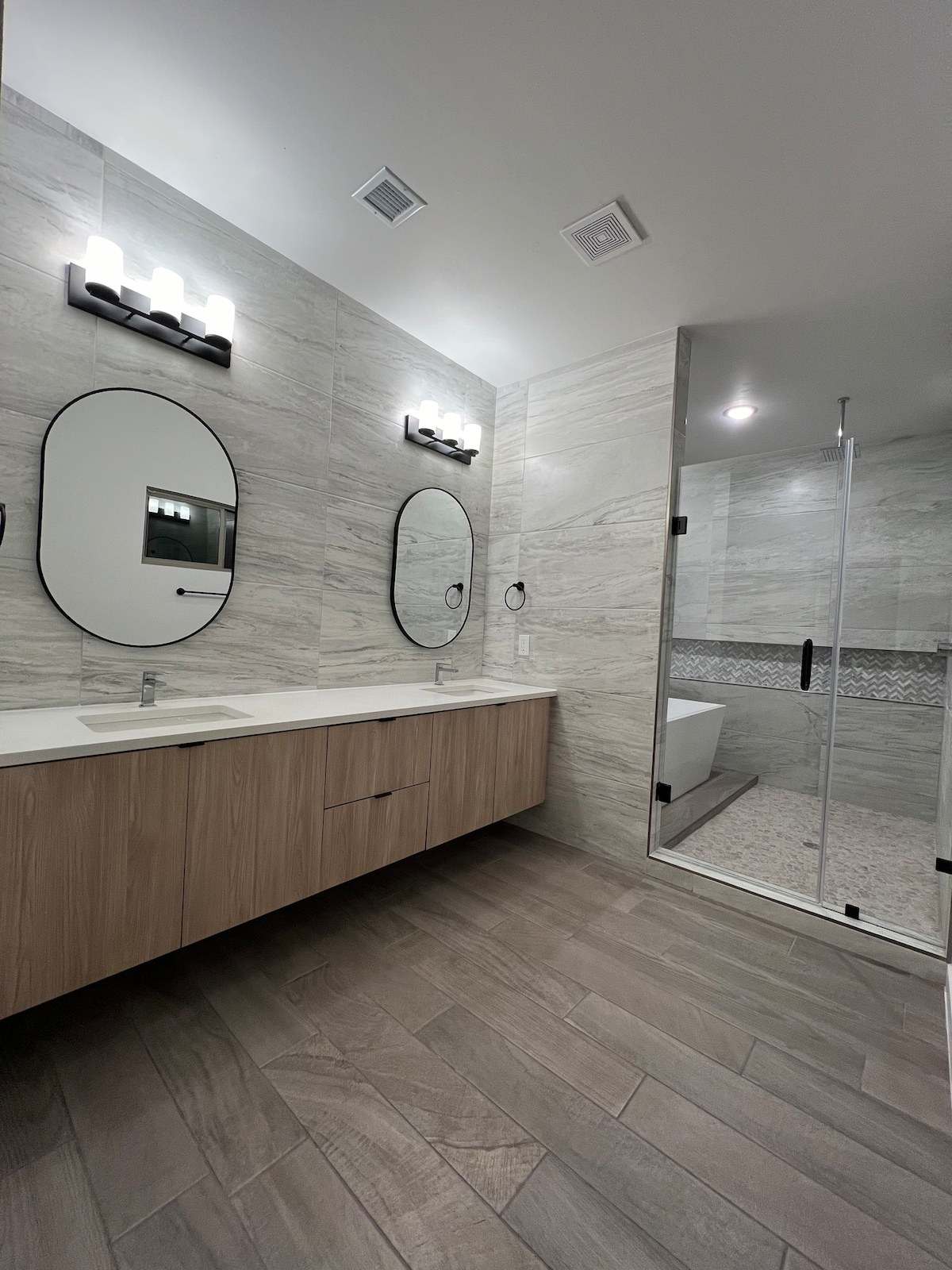 A modern bathroom featuring dual sinks with oval mirrors above each. Light wood cabinetry provides storage beneath the sinks. A glass-enclosed shower is visible in the background, with a stylish tile design on the wall. The floor is covered in large, light-colored tiles.