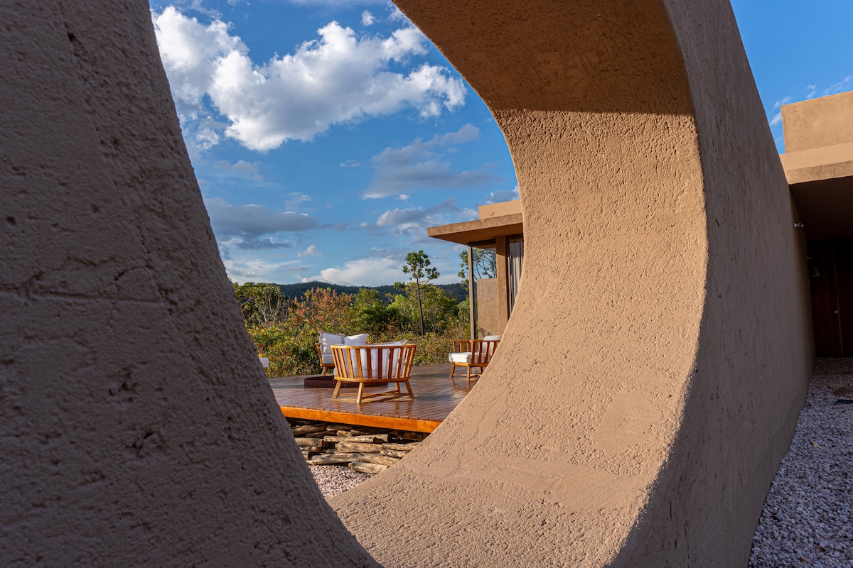 A curved architectural feature frames a view of an outdoor deck. Two wooden chairs are positioned on the deck, surrounded by greenery and distant hills under a partly cloudy sky.