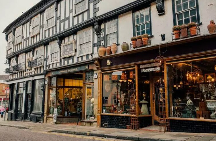 A charming street view is presented, showcasing a historic building with black and white timber-framed architecture. Vibrant shopfronts are adorned with potted plants, and various decorative items are displayed in the windows, reflecting the local culture and inviting exploration.
