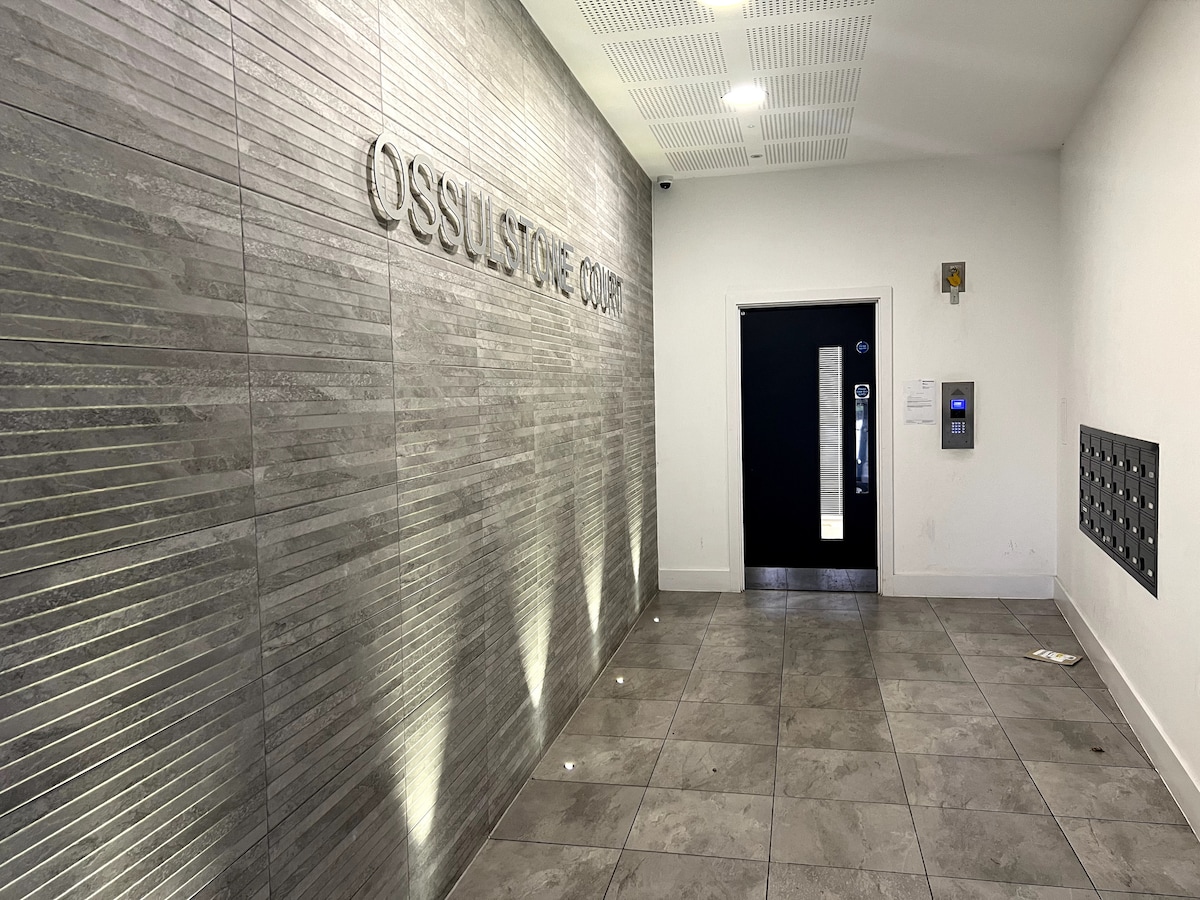 An entrance hall is presented with a textured gray wall featuring 'OSSULSTONE COURT' in bold lettering. Even lighting highlights the patterned tiles on the floor, while a black door leads to additional areas. A mail slot system is visible to the right.