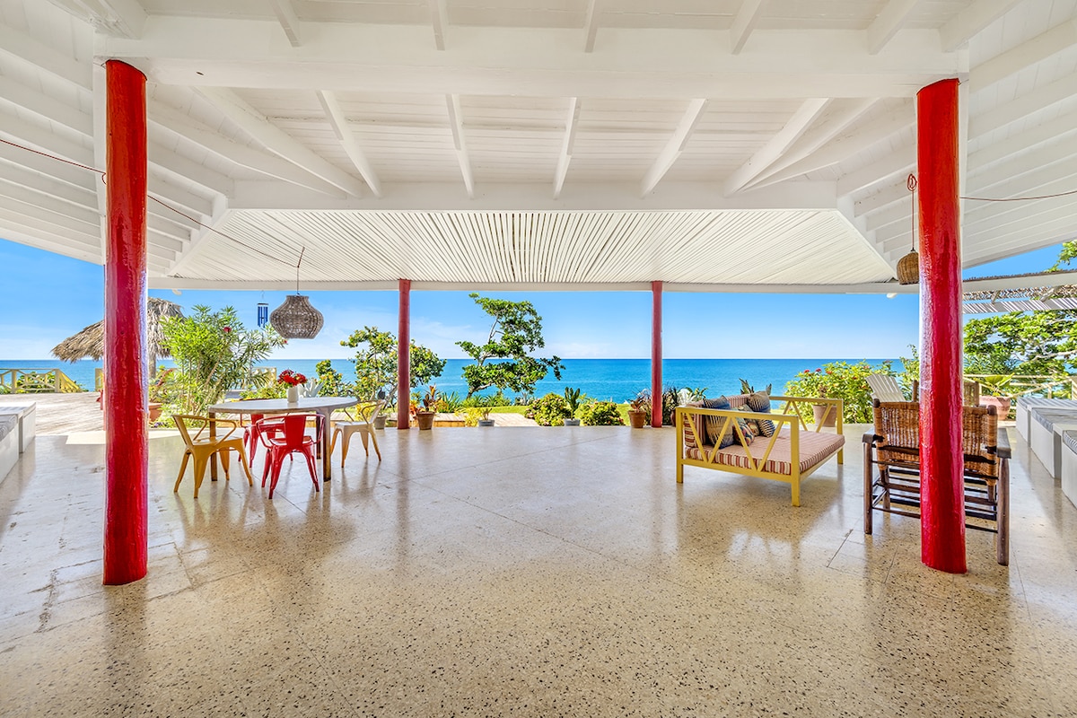 An expansive outdoor living area is depicted, featuring a large, covered space with vibrant red support beams. A dining table is situated in the foreground, accompanied by assorted chairs. The view beyond showcases the Caribbean Sea, framed by tropical greenery on the sides.
