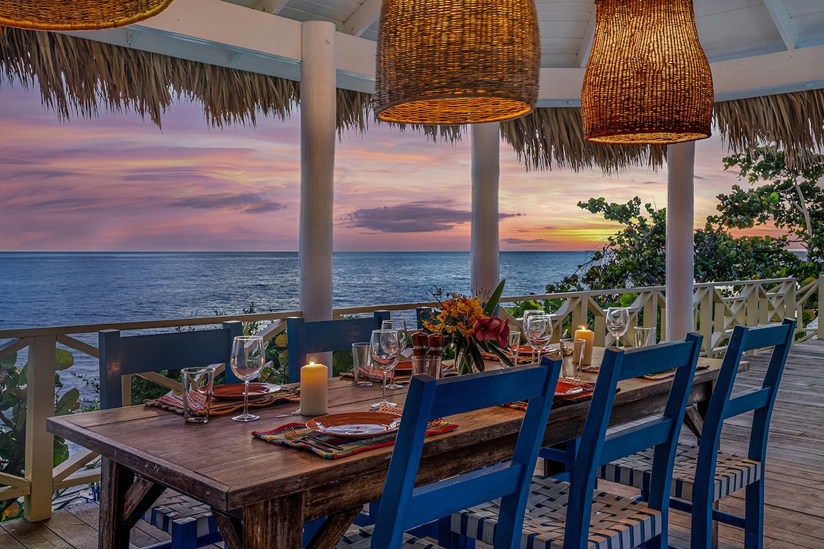 An outdoor dining area is set with a wooden table surrounded by blue chairs, featuring floral centerpieces and place settings. Warm pendant lights hang above, illuminating the scene, while the ocean and vibrant sunset create a picturesque backdrop.