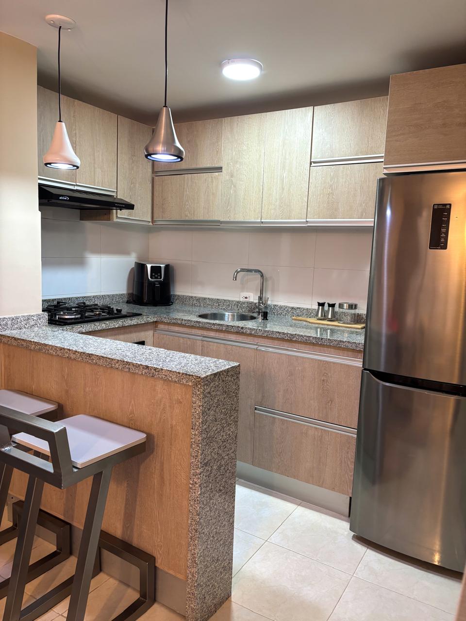 The kitchen features modern wooden cabinetry and granite countertops, equipped with a stove, sink, and refrigerator. Two pendant lights provide focused illumination above a small breakfast bar with two stools, adding functionality to the space.
