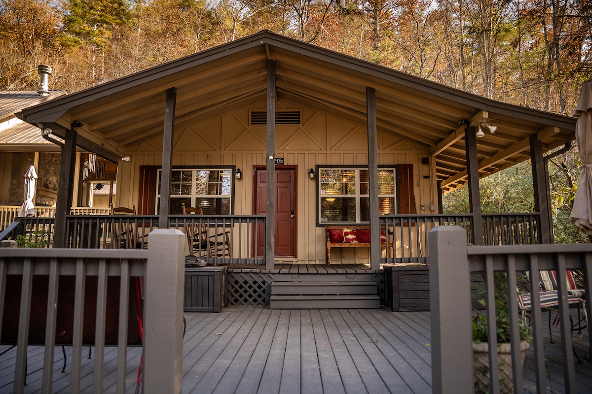 The cabin features a welcoming front porch with a slanted roof and multiple seating options. Large windows provide natural light, and the entrance is framed by a simple door. The surrounding landscape displays hints of autumn foliage.