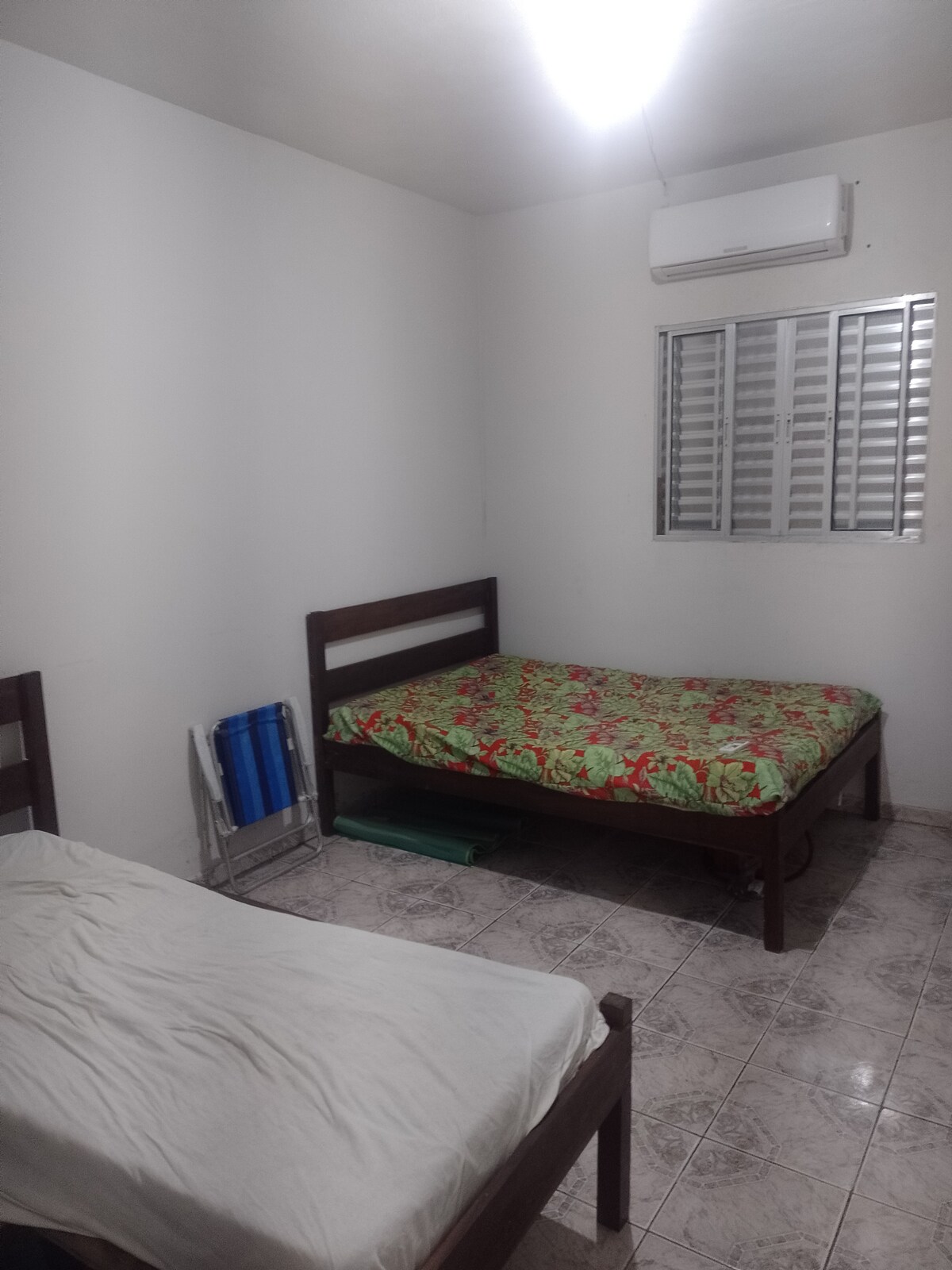 A simple bedroom is depicted, featuring two beds with colorful floral bedding. A window is present on the far wall allowing natural light. An air conditioning unit is mounted above, and a few items are arranged nearby, including a folding chair and a small green folder.