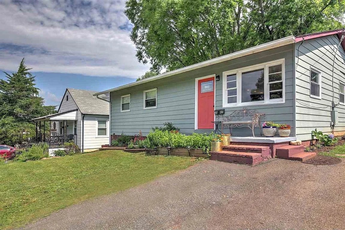 A single-story home with light blue siding is set against a clear sky. A vibrant door adds a pop of color to the entrance, which is framed by landscaped flower beds. A paved walkway leads up from the driveway, inviting visitors towards the front entrance.