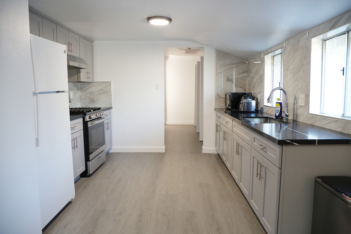 The kitchen features modern appliances, including a refrigerator and stove, alongside ample cabinetry. A long countertop provides additional workspace, complemented by a series of windows allowing natural light to fill the room. The flooring is a light, neutral color, enhancing the space's openness.