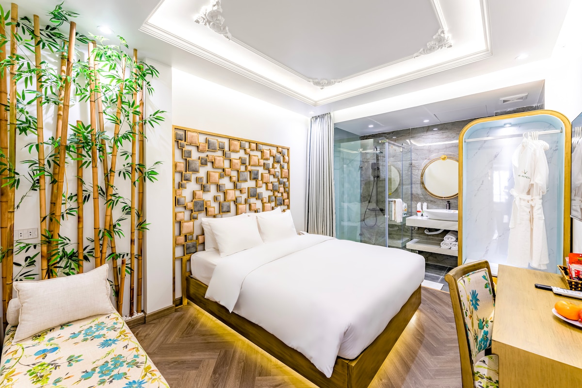 The modern room features a spacious bed against a decorative bamboo backdrop, with natural light illuminating the space. A glass-walled bathroom is visible, showcasing a shower and stylish fixtures. A small seating area is furnished with a colorful print, creating an inviting atmosphere.