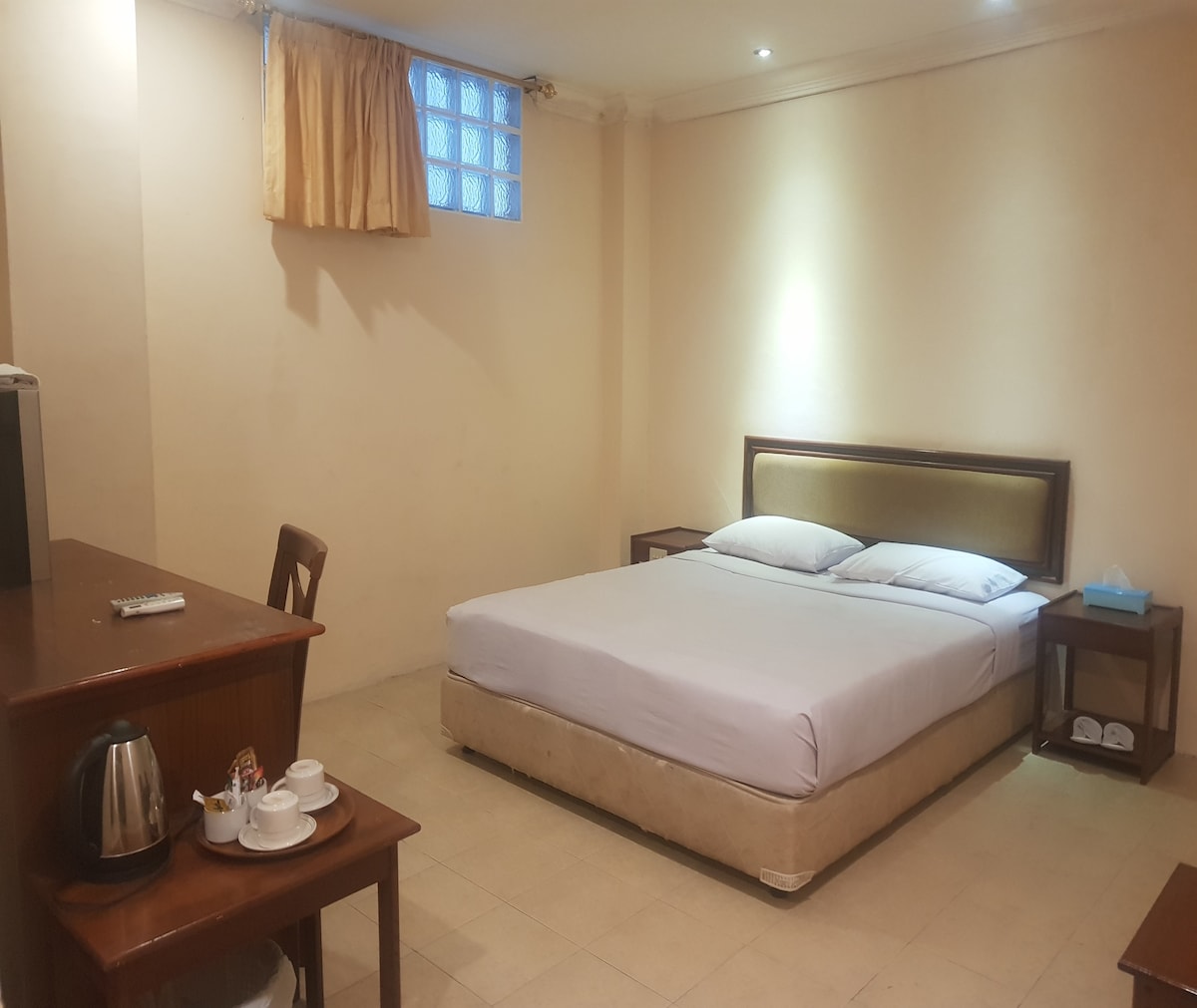 A spacious, well-lit room features a large bed with clean linens and soft pillows. A wooden desk with a chair is positioned near a kettle and tableware, creating a functional area for relaxation. Natural light filters through a window with light drapes.