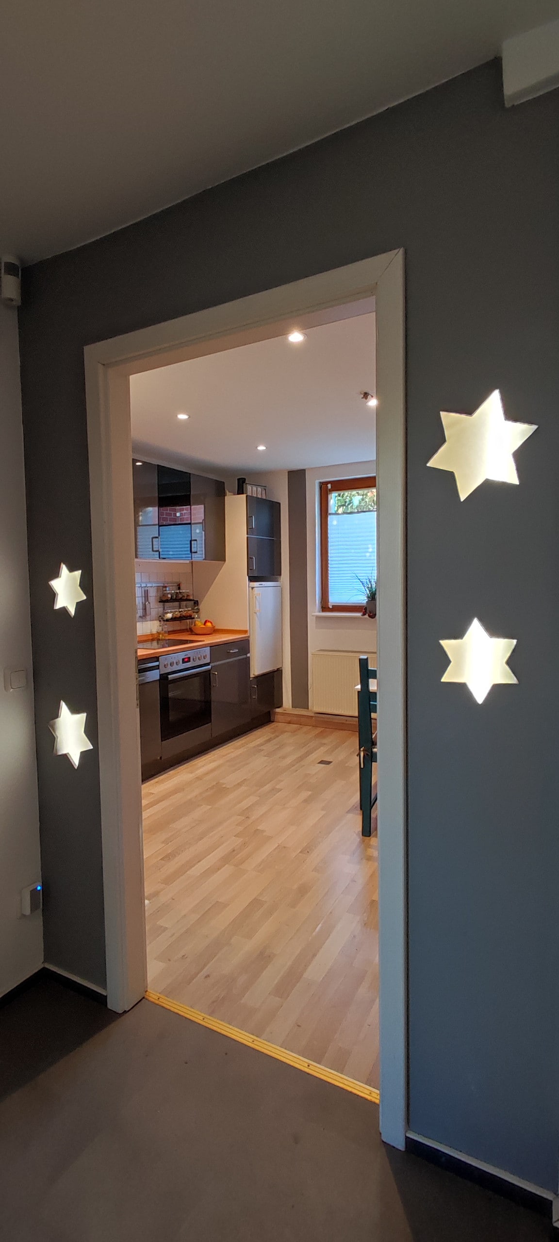 A hallway entrance is framed by star-shaped wall cutouts, leading into a bright kitchen. The kitchen features modern appliances and wooden flooring, with warm LED lighting creating a welcoming ambiance.