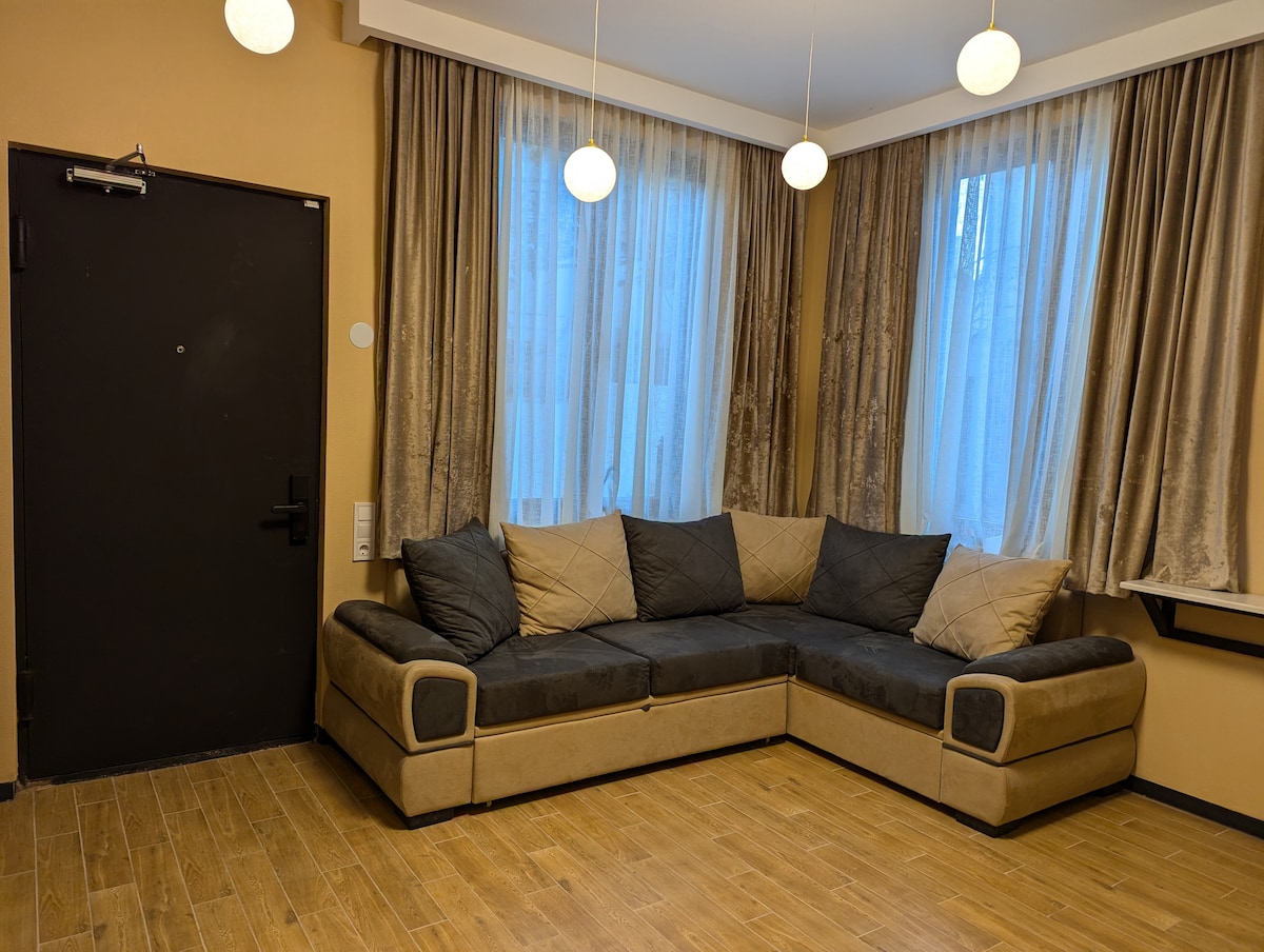 A corner seating arrangement is featured, upholstered in neutral tones, inviting relaxation. Natural light filters through sheer curtains draping over large windows. Two pendant lights hang from the ceiling, contributing to the warm ambiance of the room.