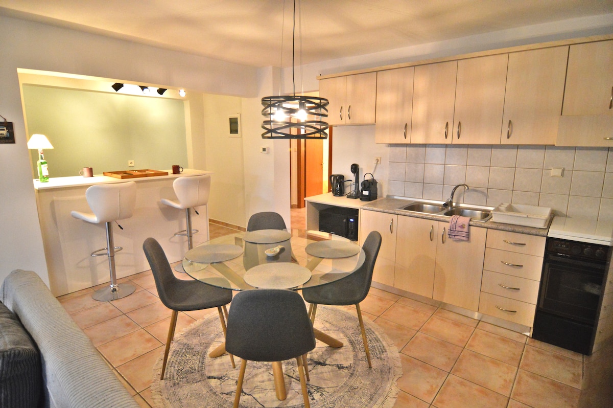 A modern kitchen area is viewable, featuring light wooden cabinetry and a countertop with appliances. A round dining table with gray chairs is centered on a circular rug, complemented by pendant lighting. A bar with two stools is visible in the background.