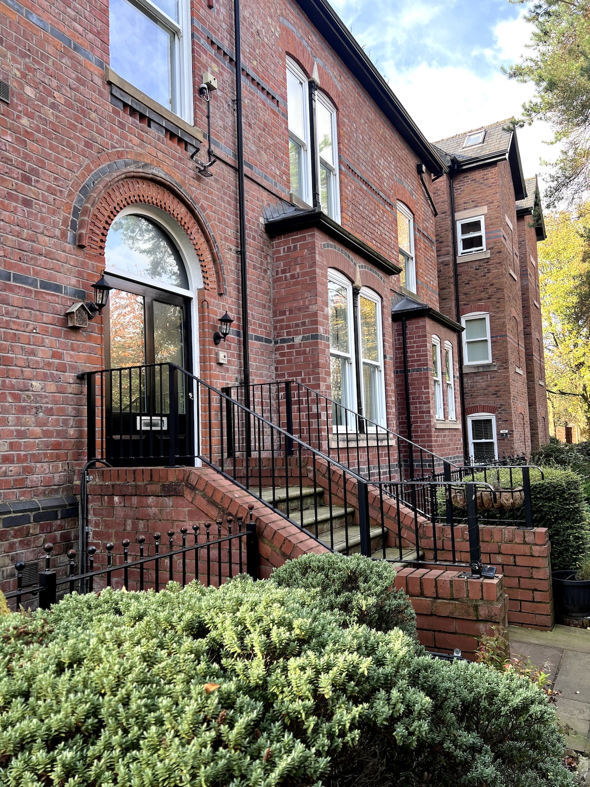 An attractive brick exterior of the apartment building is shown, featuring a set of stairs leading to a black front door framed by large windows on either side. Neatly trimmed hedges are positioned along the pathway, with trees visible in the background.