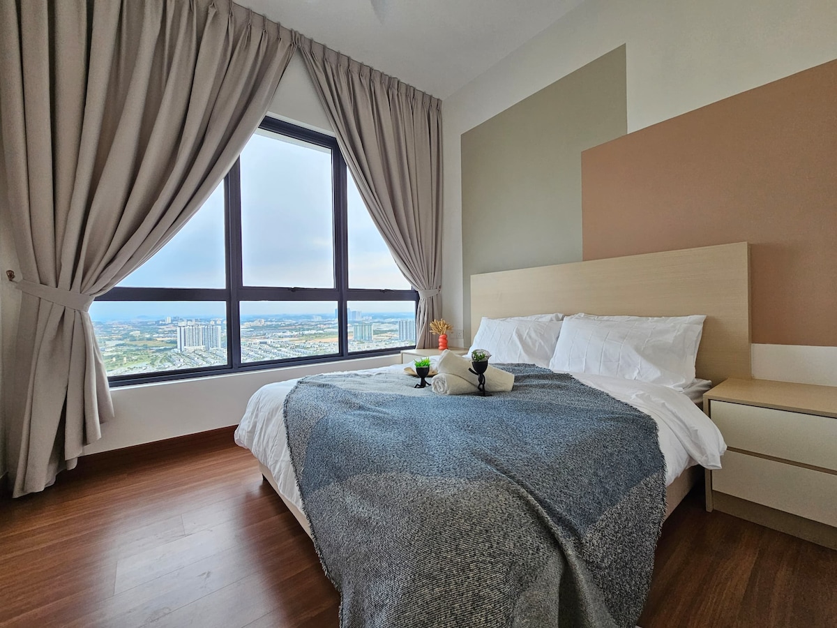 A spacious bedroom is presented with a large window offering expansive views. A comfortable bed is dressed in white linens, accompanied by coordinating throw pillows. A textured blanket is placed across the bed. Soft curtains frame the window, enhancing the room's inviting nature.