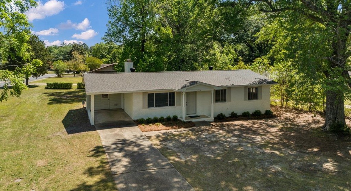 A single-story home is surrounded by lush green trees and a well-maintained lawn. The inviting front porch features a small set of steps, while a spacious driveway runs alongside the house. Natural sunlight illuminates the exterior, creating a warm and inviting look.