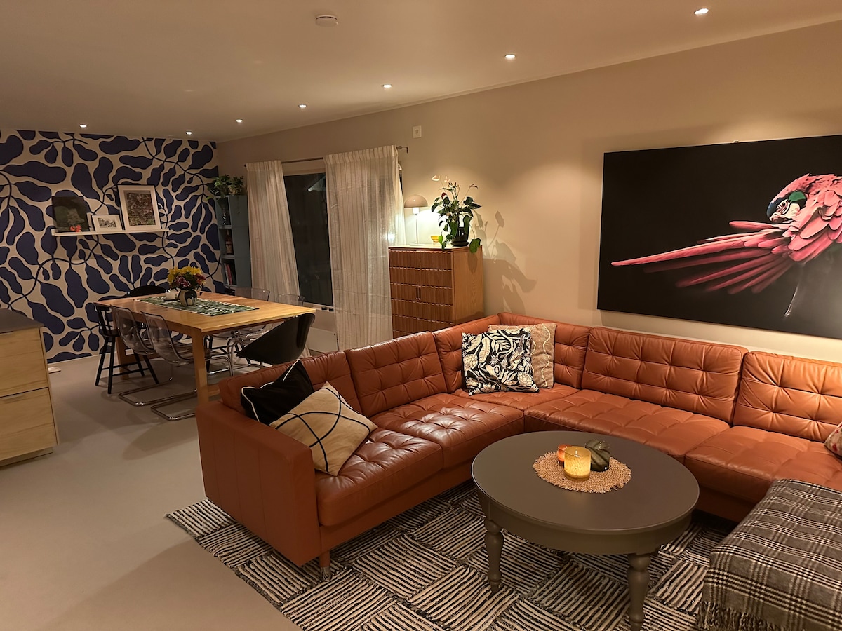 A spacious living area showcases a leather sectional couch, accented by patterned cushions. A round coffee table sits at its center. The background reveals a dining area with a wooden table and chairs, complemented by a colorful wall mural and illuminated by soft lighting.