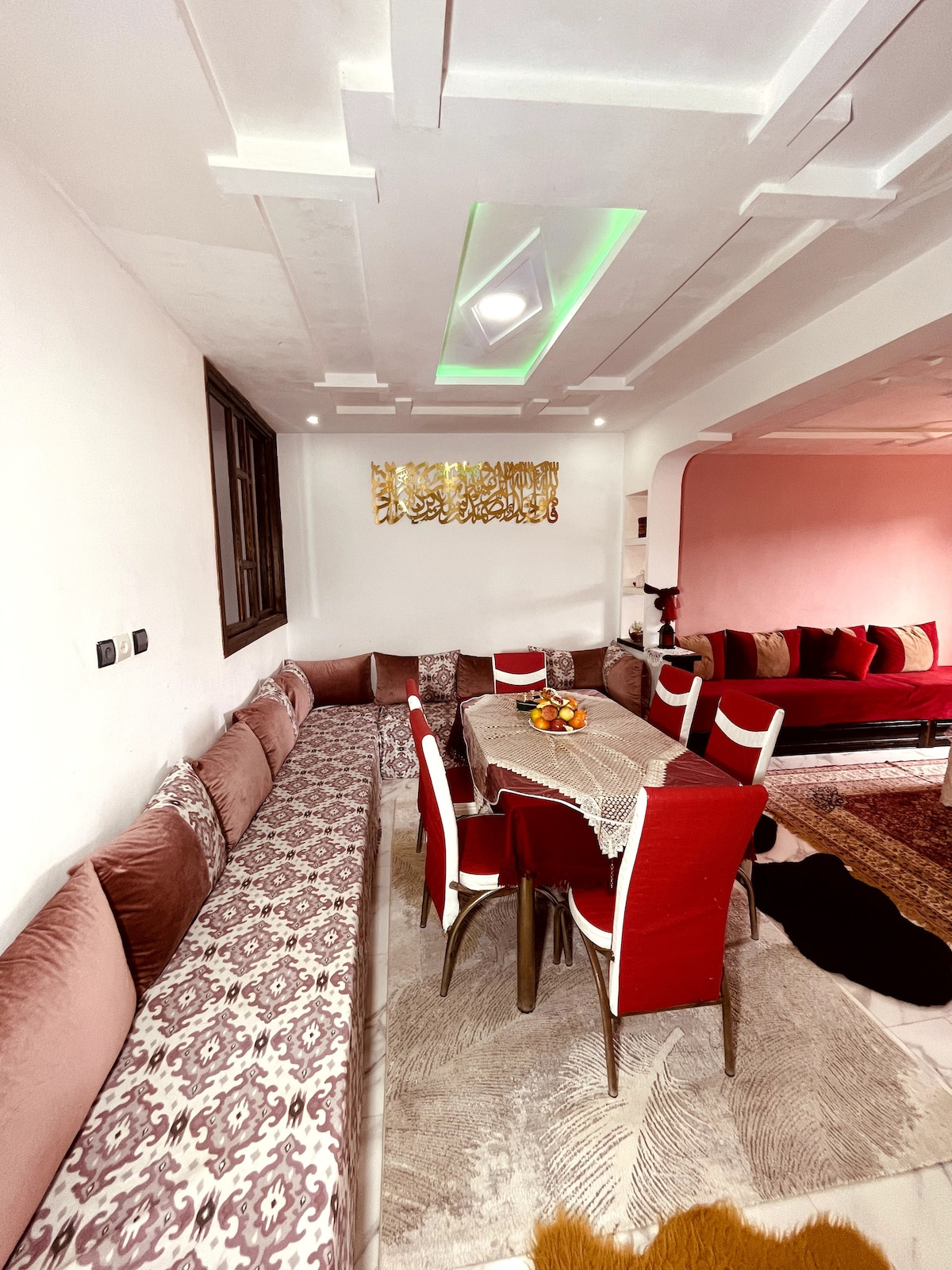 A dining area features a table surrounded by six red chairs. A plush, L-shaped sofa with decorative cushions lines the wall. Decorative artwork is displayed overhead, with soft lighting enhancing the cozy atmosphere. A patterned rug adds texture to the space.
