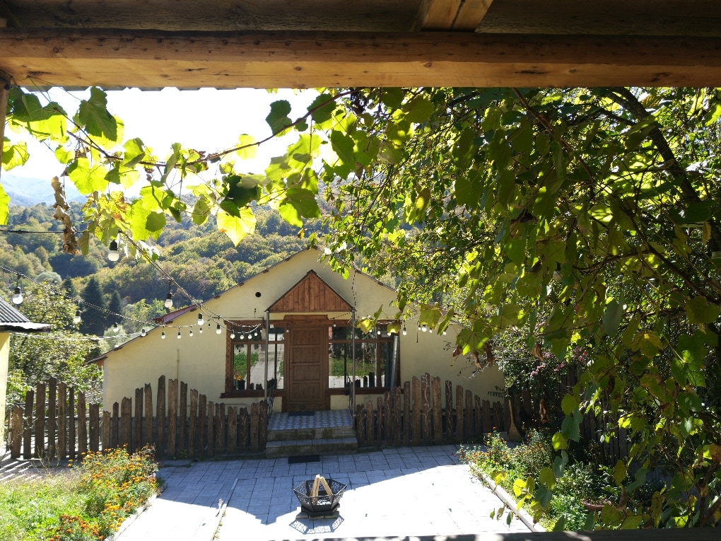 The image captures a charming house framed by a wooden porch, surrounded by greenery. The structure features a wooden front door and a well-maintained garden, with a pathway leading to the entrance. String lights are visible, enhancing the inviting outdoor space.