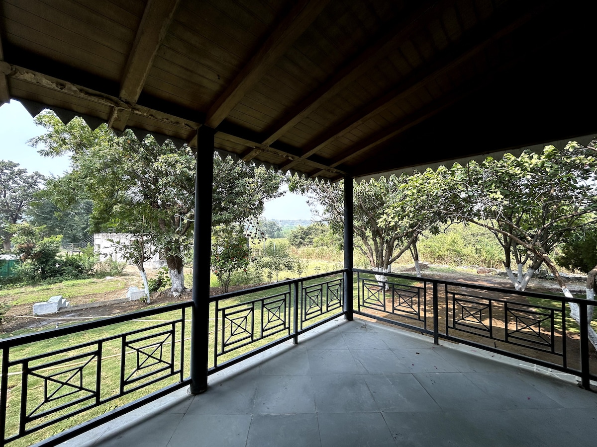 A spacious veranda offers a view of the surrounding farmland, featuring lush greenery and trees. The wooden ceiling enhances the area, while the black wrought-iron railings frame the outdoor landscape, creating a peaceful space for relaxation.