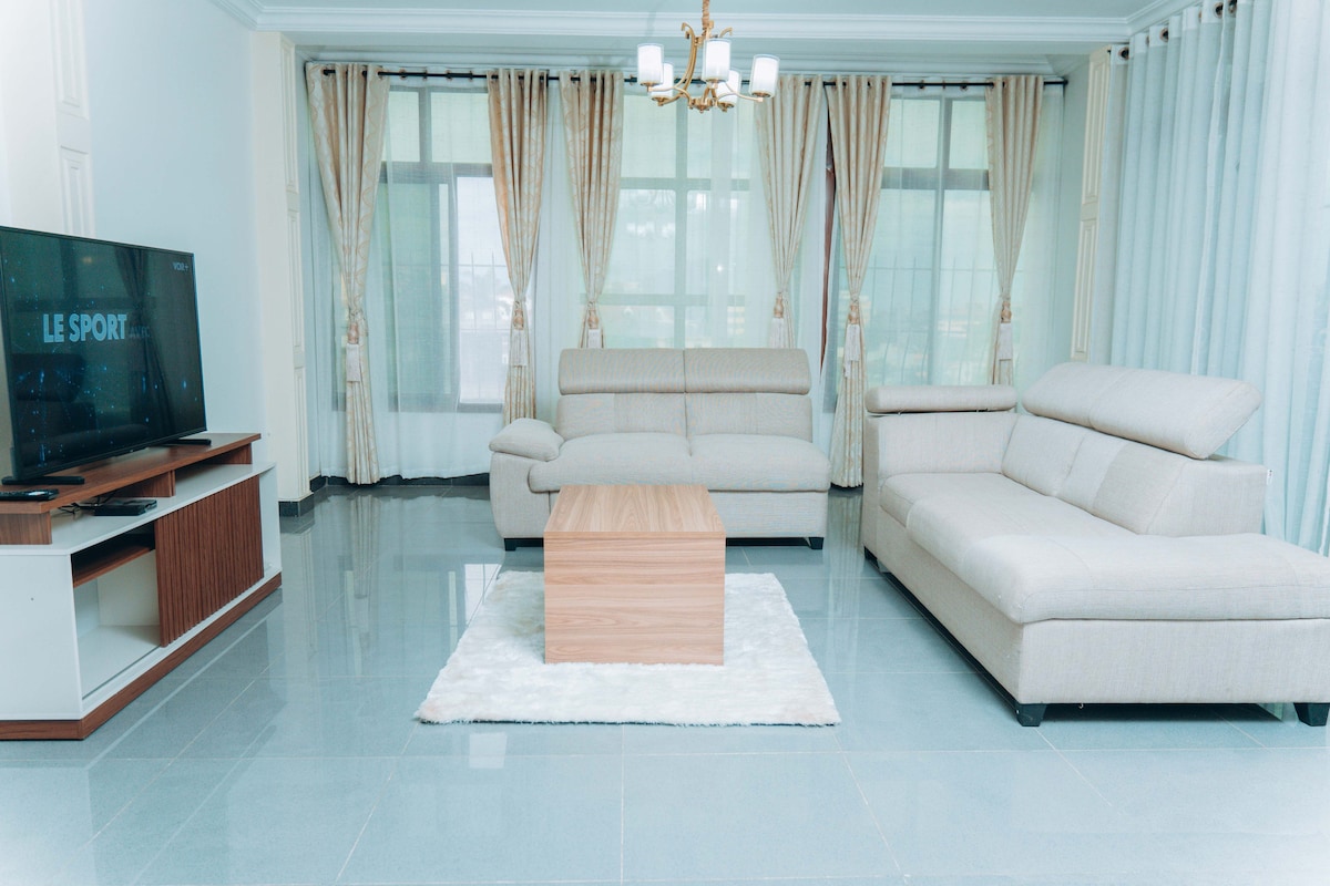 A spacious living area features large windows draped with light, flowing curtains. A soft, modern sectional couch is complemented by a matching armchair. A wooden coffee table rests on a soft area rug, while a flat-screen television is mounted against the wall.