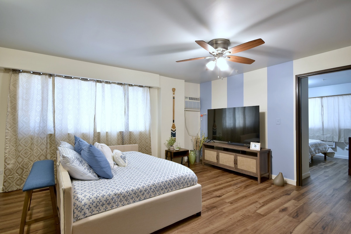 The bedroom is furnished with a spacious bed, light bedding, and decorative pillows. A large flat-screen smart TV is positioned on a media console. Natural light filters through sheer curtains, and a ceiling fan adds to the room's comfort.