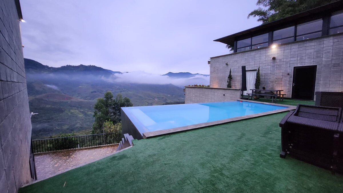 An expansive outdoor area showcases a serene swimming pool bordered by lush greenery and mountain views. The clear water reflects the sky, and seating is arranged nearby for relaxation. Low clouds rest over the distant hills, creating a tranquil atmosphere.