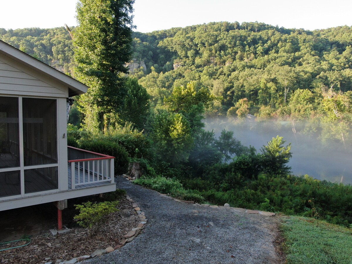 A scenic view captures the home with a screened porch, nestled beside a winding gravel pathway. Lush greenery and trees rise in the background, partially shrouded in morning mist, while the tranquil waters of the Little Red River reflect the surrounding natural beauty.
