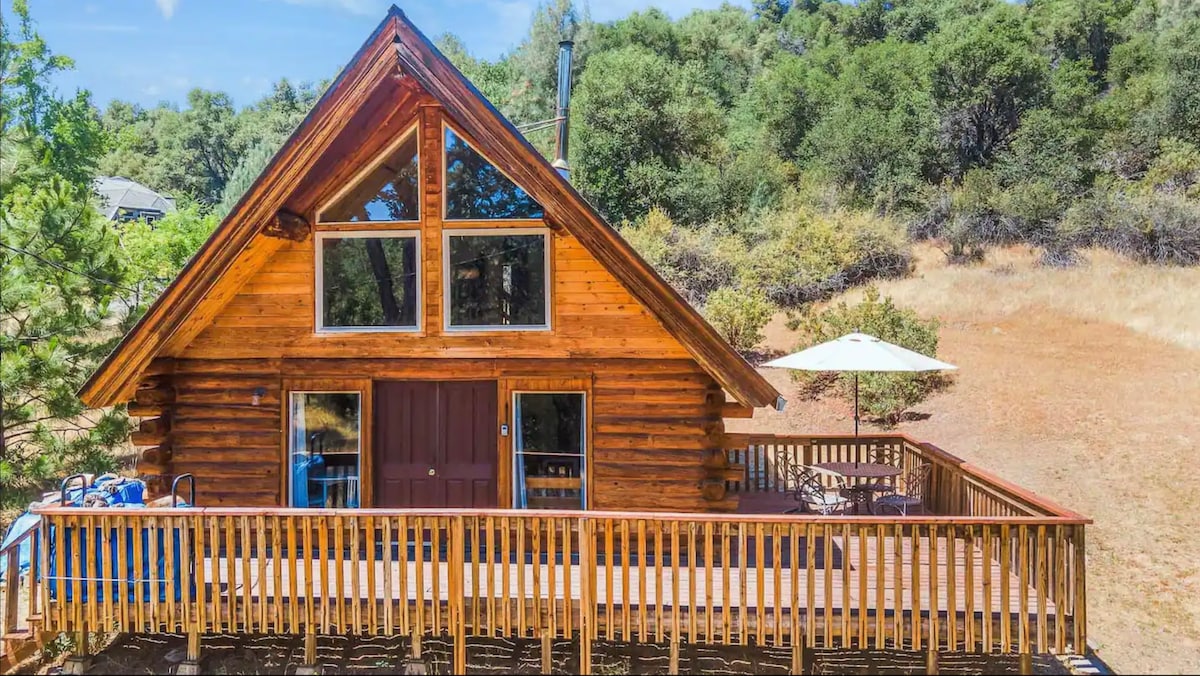 The log cabin features a sloped roof with large windows allowing natural light to illuminate the space. A spacious deck wraps around the cabin, furnished with a table and chairs, surrounded by natural landscaping and mature trees.