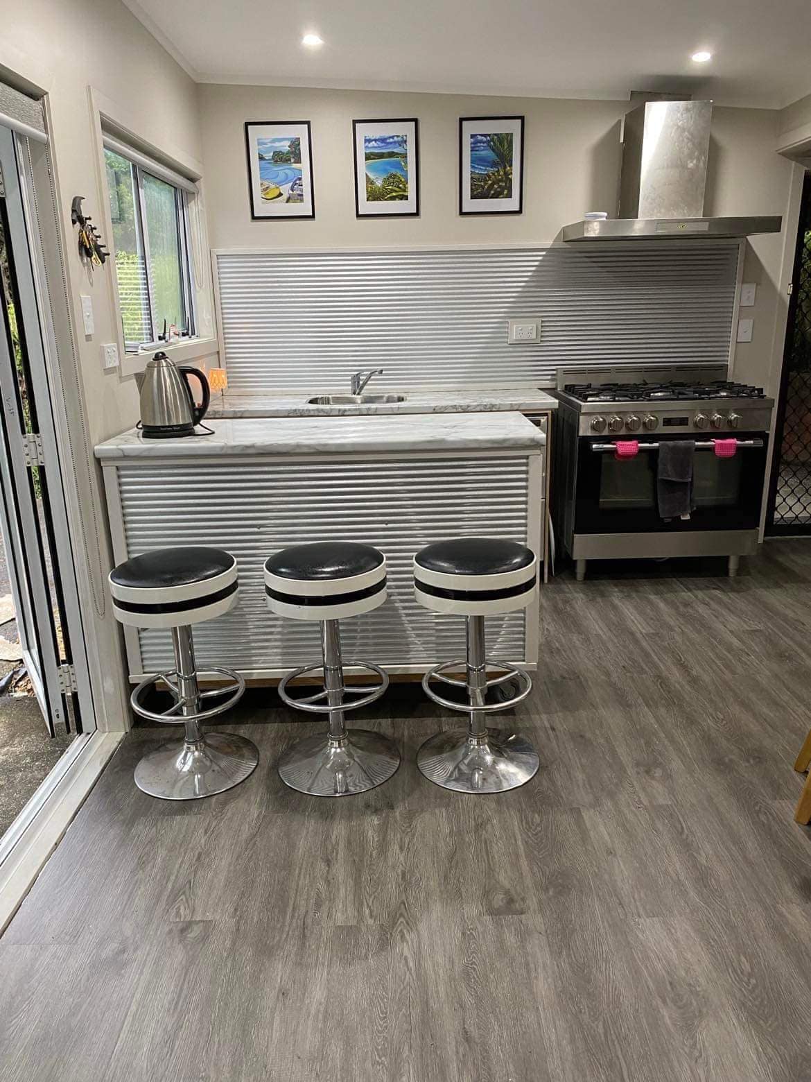 A modern kitchen is presented, featuring a marble-topped island with three black and silver bar stools. The kitchen includes stainless steel appliances and a large gas stove. Framed art depicting landscapes adorns the wall above the sink, which is positioned under a window allowing light to enter.
