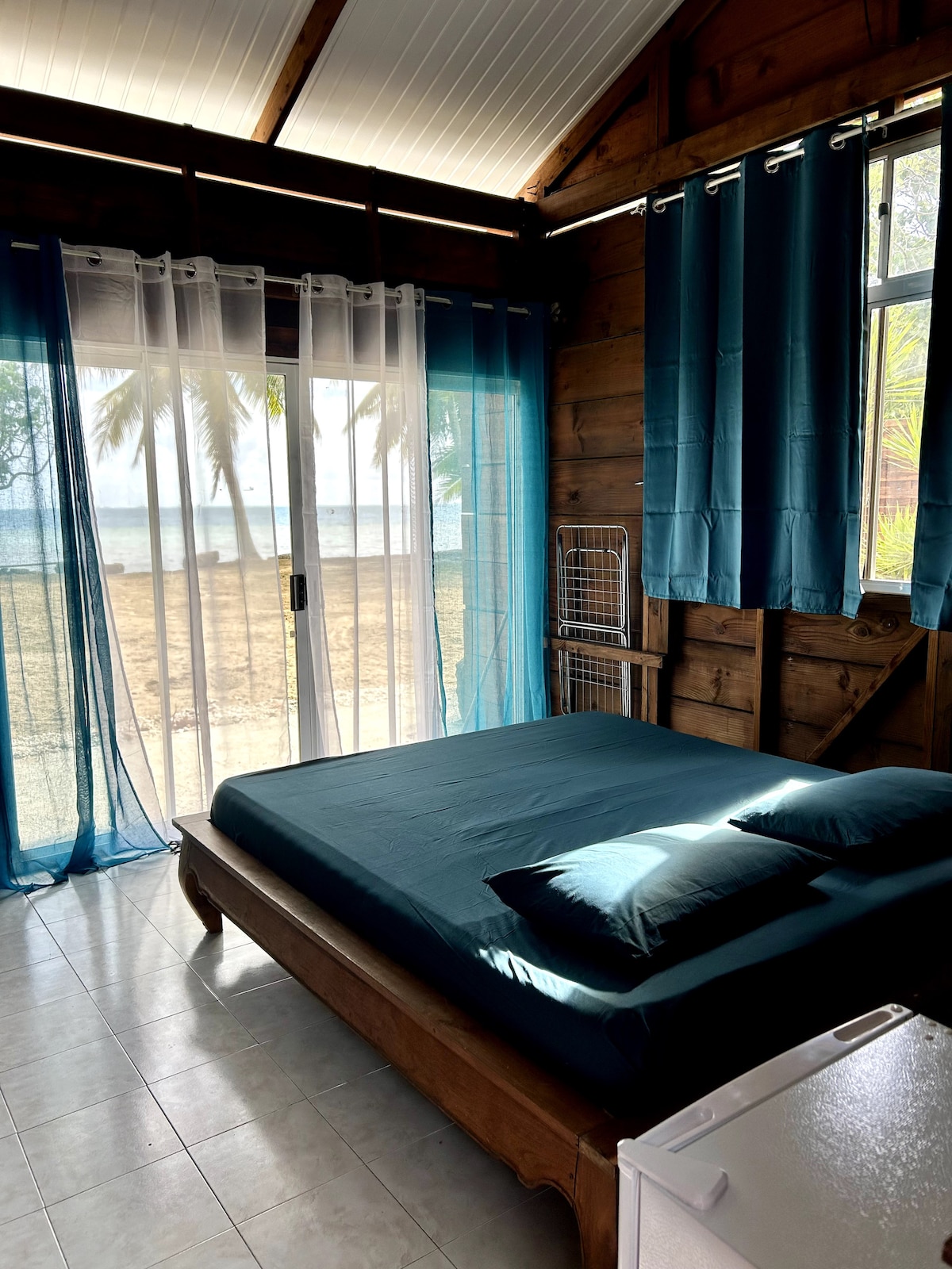 A bright sleeping area features a spacious bed with teal bedding and two matching pillows. Sheer curtains allow natural light to filter in, framing a view of the beach outside. Wooden walls add warmth to the serene environment.