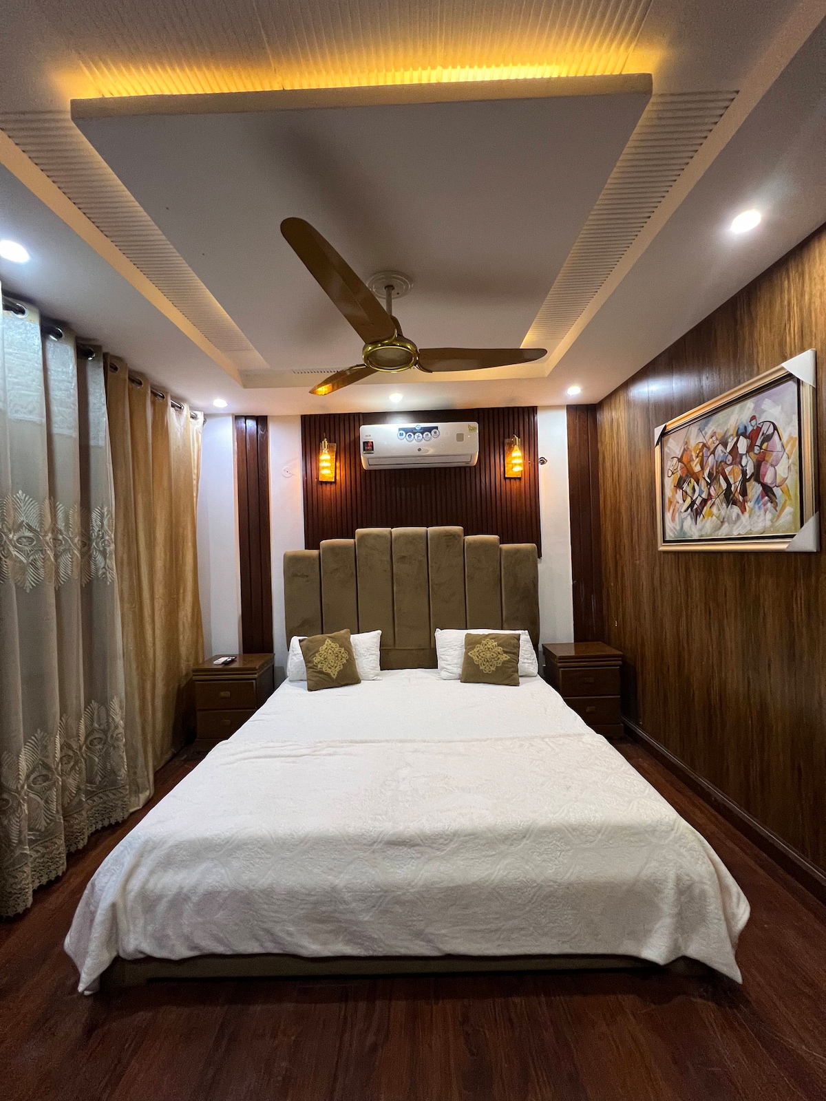 A serene bedroom is showcased, featuring a spacious bed with a light-colored cover and coordinating decorative pillows. Warm wooden paneling complements the room's modern decor, while a ceiling fan and soft lighting enhance the ambiance. A large window adorned with elegant curtains allows for natural light.