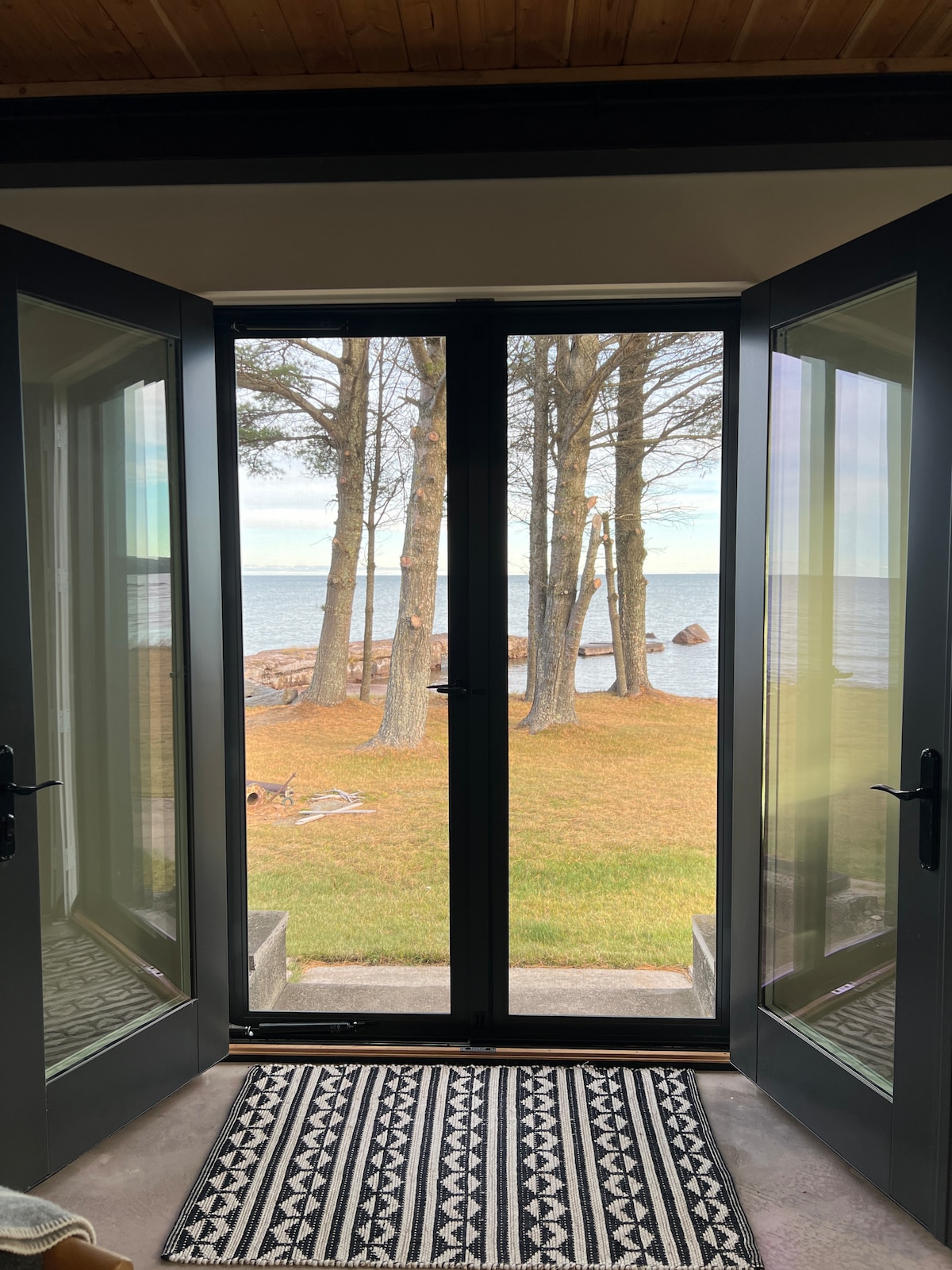 A view through double glass doors reveals a serene landscape of Lake Superior. Framed by tall trees and grassy areas, the shoreline includes distant rocky formations. A patterned area rug is visible in the foreground, adding texture to the indoor space.