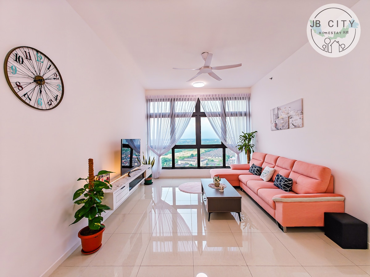 A well-lit living area showcases a large window with scenic views. A comfortable orange sofa is arranged around a low coffee table, accompanied by a potted plant. A wall-mounted television is positioned opposite the seating, and a large wall clock adds a practical touch.