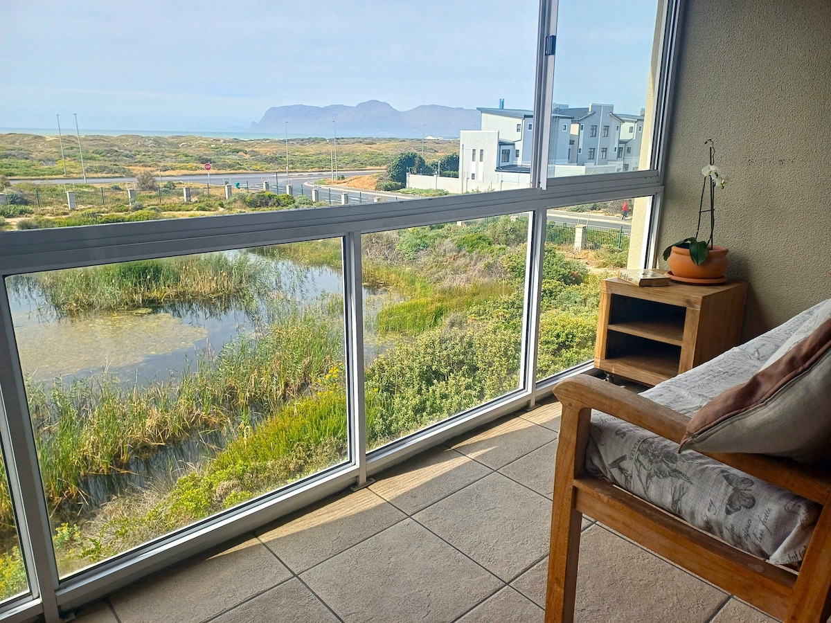 A seated area featuring wooden furniture is positioned beside expansive glass windows, offering a view of a wetland bird sanctuary and scenic landscape. Natural light illuminates the space, highlighting greenery and the distant coastal hills through the glass.