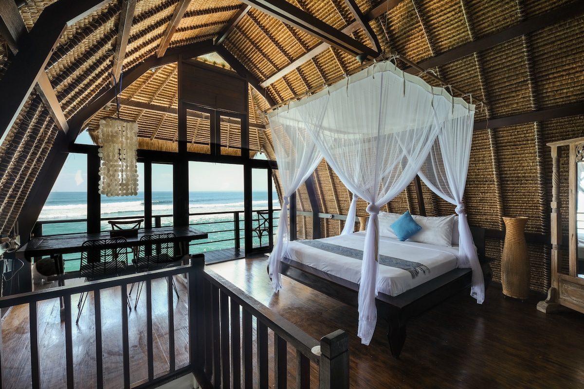 The spacious bedroom features a king-size bed with a mosquito net, positioned to offer ocean views through large windows. A small dining area with a table and chairs complements the room, while wooden accents and woven ceilings enhance the traditional aesthetic.