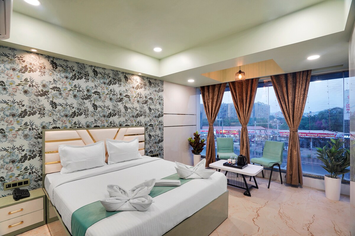 A bright and inviting bedroom features a spacious bed with neatly arranged towels resembling decorative swans. The floral wallpaper complements the soft green accents. A large window offers a view of the outdoors, framed by elegant curtains, while a small table and chairs are positioned nearby.