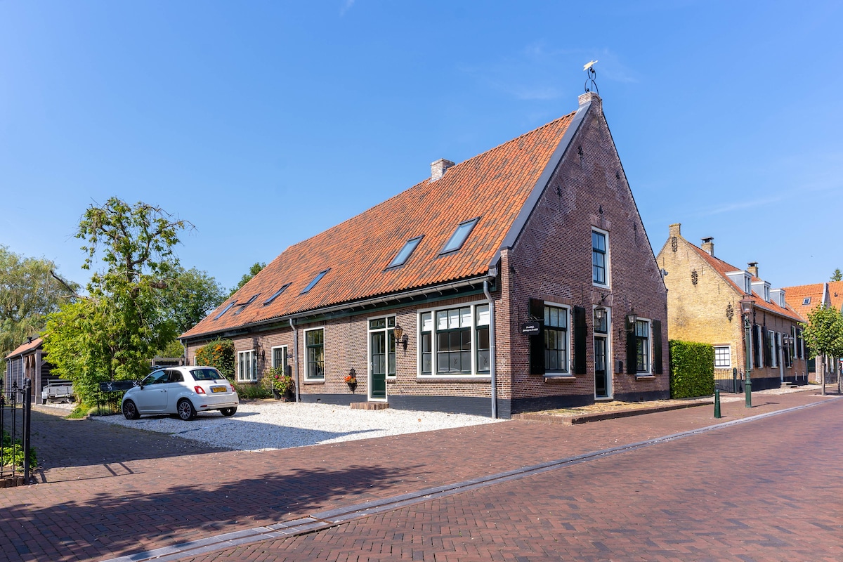 The exterior of a renovated historical farmhouse is presented, showcasing a traditional brick façade and a sloped red-tile roof. A well-maintained gravel driveway allows for parking, with a nearby tree providing greenery against a clear blue sky.