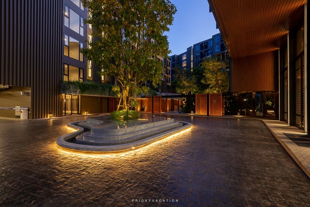 An outdoor area features a serene water feature surrounded by stone pathways and softly illuminated by built-in lighting. Lush trees and greenery create a calming environment, with modern buildings framing the space in the background.