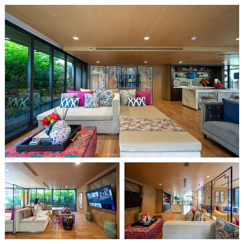 A spacious living area features large windows allowing natural light and views of greenery. Seating includes a light-colored sofa adorned with patterned cushions. A decorative table with a floral arrangement adds a touch of color. A smart TV is mounted on the wall across from the seating.