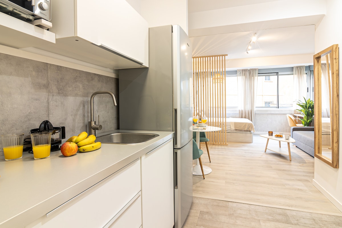 A well-equipped kitchen presents a modern design, with a stainless steel refrigerator and a sink. Fresh fruits and two glasses of orange juice are placed on the countertop. The open layout allows views into the living area, featuring light-colored furnishings and natural light from the window.