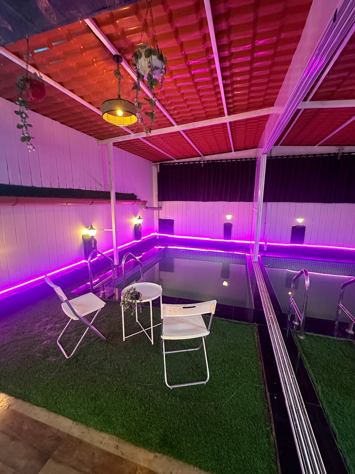 An intimate area is highlighted by a private plunge pool, surrounded by artificial grass. Two white chairs and a small round table are positioned nearby, with soft ambient lighting from overhead fixtures enhancing the secluded atmosphere.