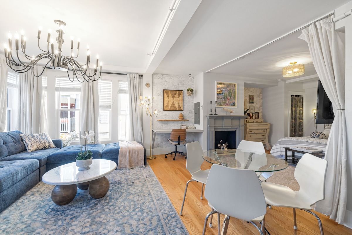 A spacious living area is highlighted by a large sectional sofa and a round coffee table. Natural light filters through multiple windows, creating a bright environment. A dining table with modern chairs is positioned nearby, alongside a fireplace and decorative art pieces on the wall.