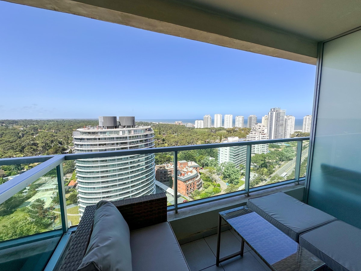 A private balcony offers panoramic views of the surrounding landscape and coastline. The space features a comfortable seating area with stylish furniture, including a sleek table and lounge chairs, providing an ideal spot to unwind while enjoying the scenery.