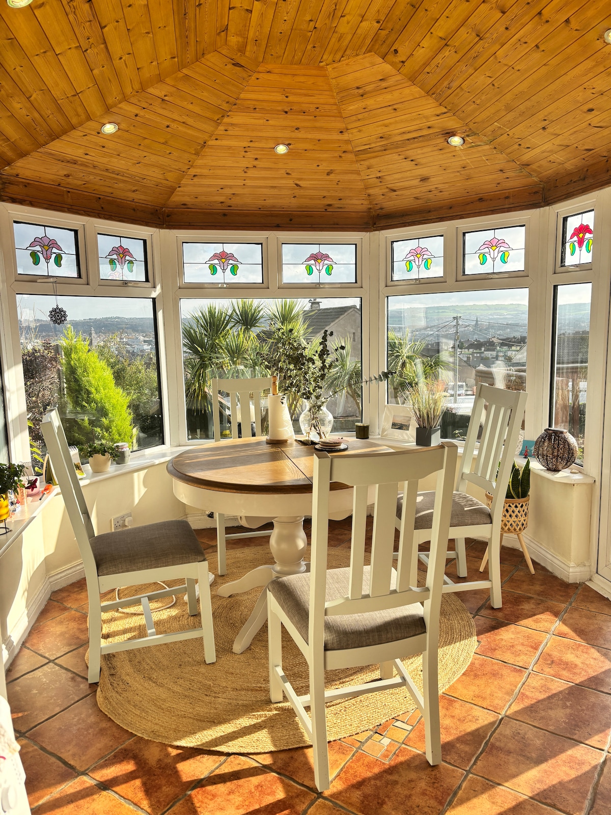 A bright conservatory features a circular wooden table surrounded by four light-colored chairs. Large windows adorned with stained glass allow natural light to fill the space, showcasing views of the surrounding greenery and hills. A woven rug enhances the inviting feel of the area.