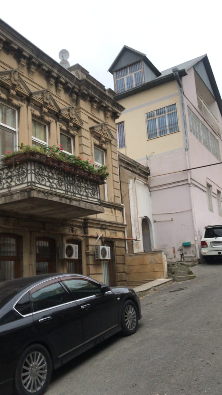 A street view features a harmonious blend of architectural styles, with an ornate building showcasing wrought iron balconies and greenery. Adjacent, a modern structure with large windows rises, and a parked vehicle is visible in the foreground against the cobblestone road.