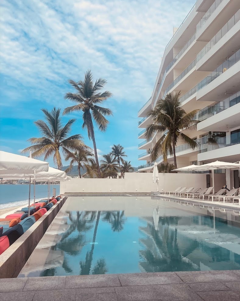 A serene infinity pool is framed by palm trees and colorful lounge chairs. The clear water reflects the bright sky and the modern building facade, creating a refreshing atmosphere for relaxation. Umbrellas provide shade around the pool area.