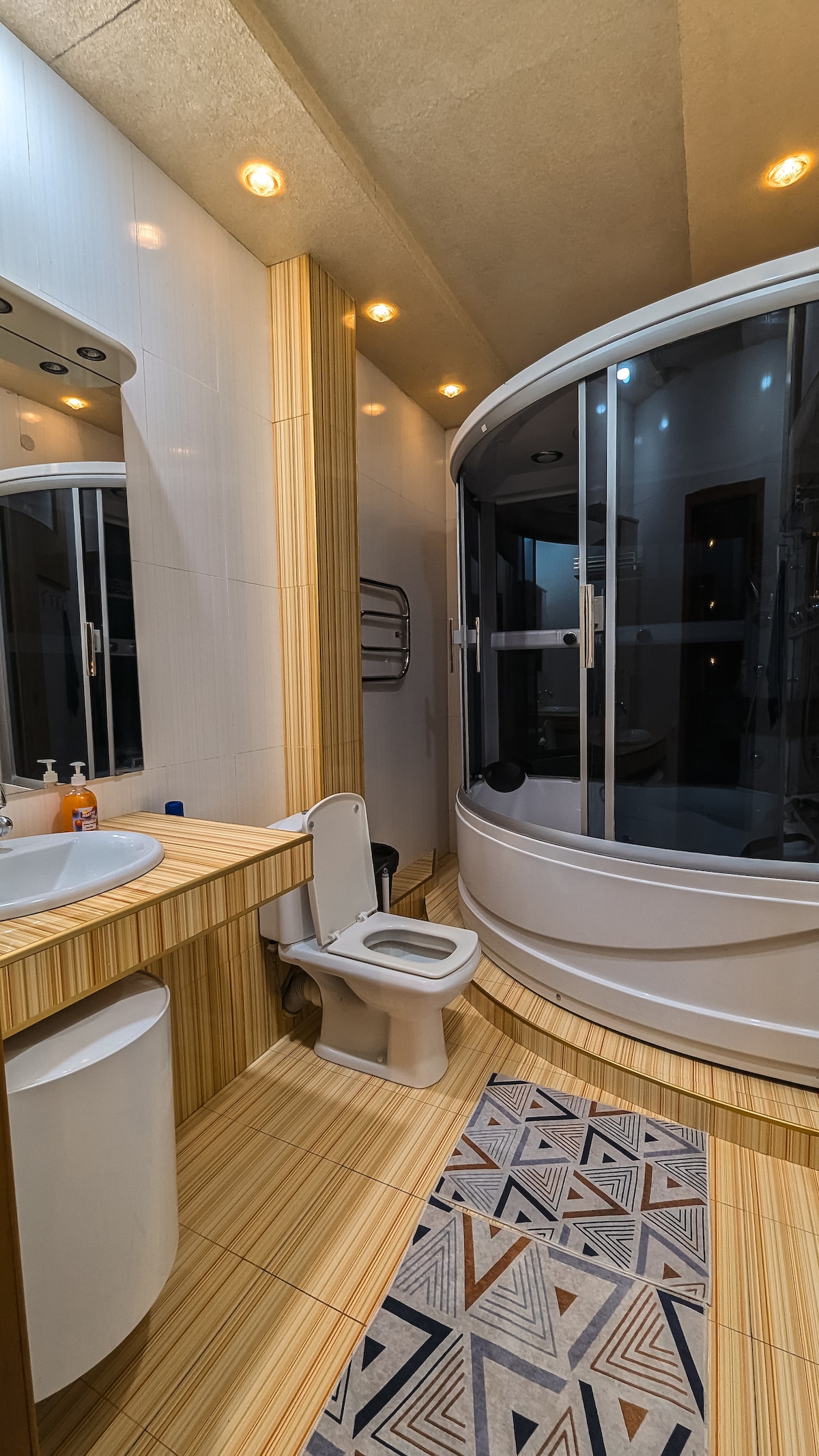 The bathroom features a modern design with light wood accents and sleek finishes. A curved shower enclosure complements a spacious vanity with a round sink. The patterned rug adds visual interest, and large mirrors enhance the sense of space and brightness.