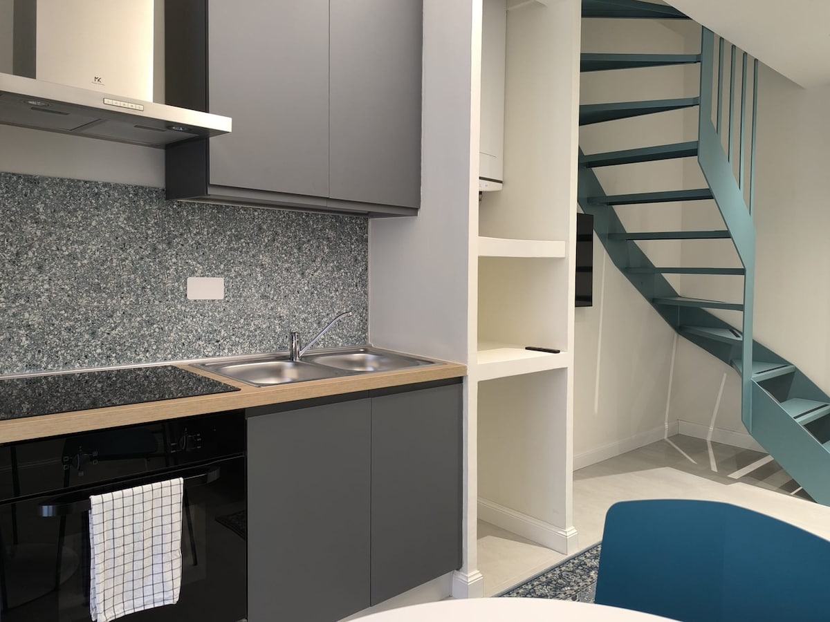 The modern kitchen area features sleek cabinetry with a dual-basin sink and an electric stove. A textured blue backsplash adds visual interest. In the background, a spiral staircase in soft teal leads to the upper level, enhancing the overall design of the space.